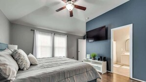 Ein Schlafzimmer mit Bett, Deckenventilator, an der Wand montiertem Fernseher und einer Tür zu einem Badezimmer.