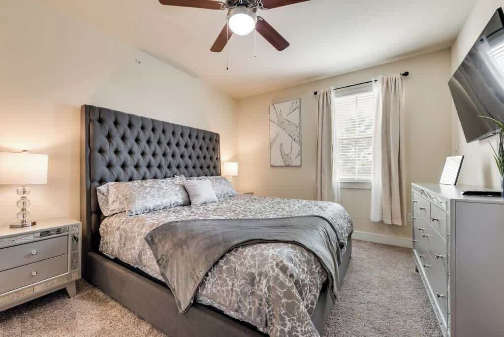 Een slaapkamer met een gestoffeerd hoofdeinde, een bed met beddengoed met een patroon, een nachtkastje met een lamp, een dressoir en een plafondventilator.
