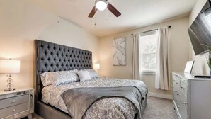 A bedroom with a tufted headboard, a bed with patterned bedding, a nightstand with a lamp, a dresser, and a ceiling fan. - 3BD  Pool Table Dallas Dallas DAL212 (Dallas)