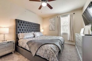 Een slaapkamer met een gestoffeerd hoofdeinde, een bed met beddengoed met een patroon, een nachtkastje met een lamp, een dressoir en een plafondventilator.