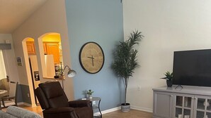 En stue med en grå hjørnesofa, en brun lænestol, et fjernsyn på et gråt skab, et stort vægur og en potteplante.