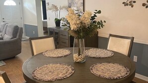 En spiseplads med et rundt træbord, seks stole, en vase med blomster og en lysekrone.