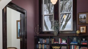 Quarto com piso de madeira, estante cheia de livros, janela com vista para árvores e luminária pendente.
