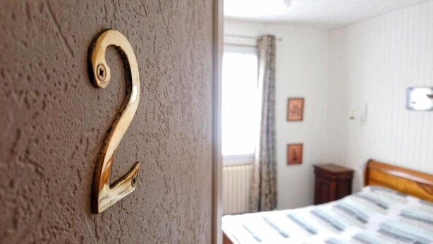 Una habitación con el número 2 de madera en la pared, una cama con cubrecama a rayas, una ventana con cortinas y una mesita de noche pequeña.