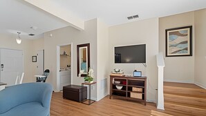 Sala de estar com sofá azul, piso de madeira, televisão em um suporte, um espelho e um quadro na parede.