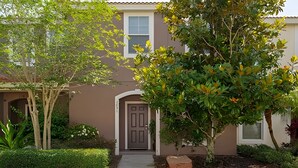 Uma casa de dois andares com fachada marrom, porta da frente e um pequeno jardim com diversas plantas e uma árvore.