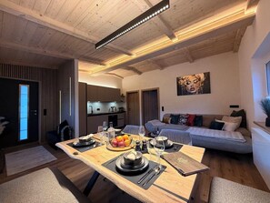 Ein Essbereich mit einem Holztisch, gedeckt für eine Mahlzeit, einer Couch im Hintergrund und einer durchgehenden Tür sichtbaren Küche.