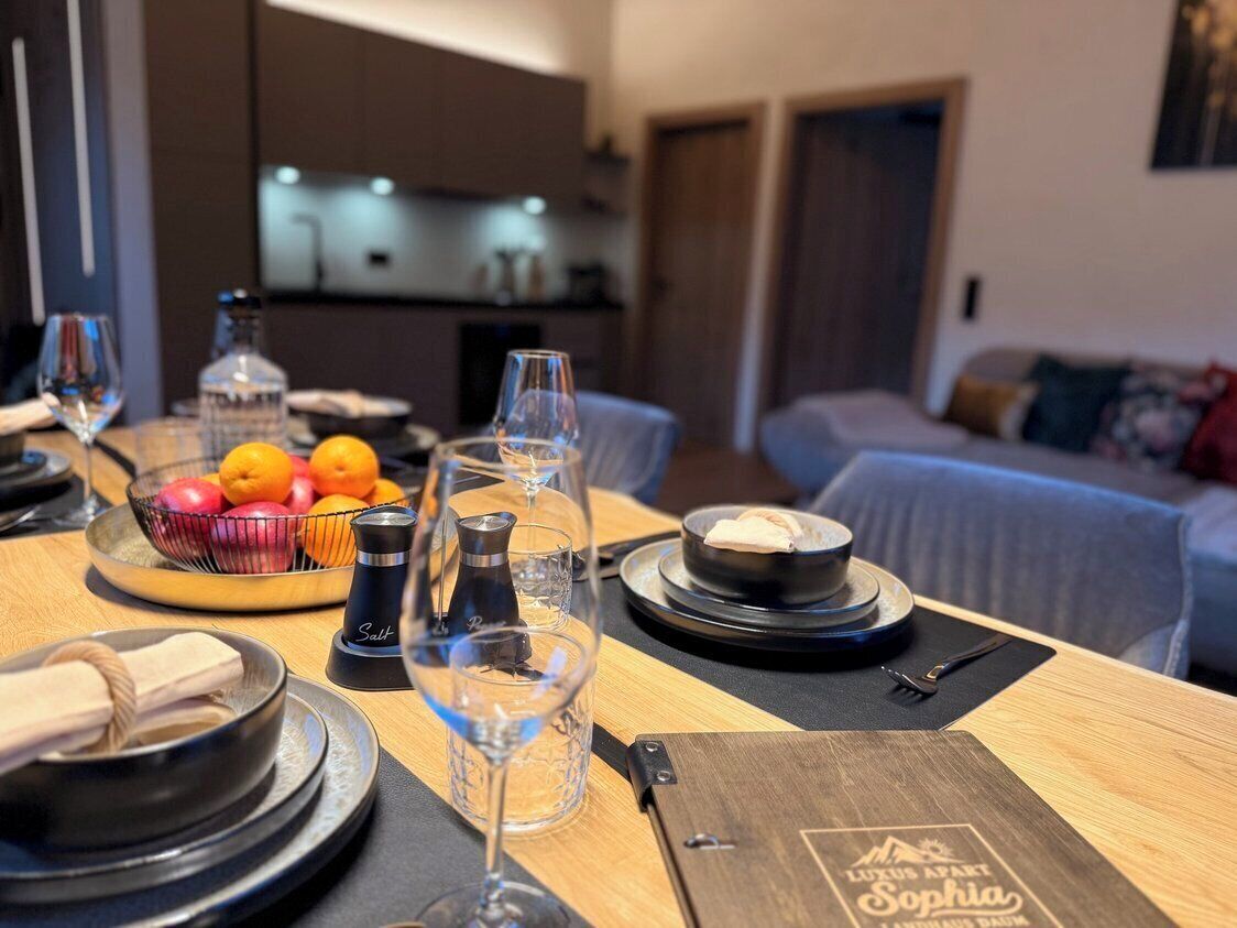 A dining table set with plates, bowls, glasses, and a bottle, with a sofa and a bed in the background.