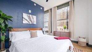 Une chambre d’hôtel moderne avec un grand lit, une vue sur les immeubles, une plante en pot et une fenêtre avec des rideaux.