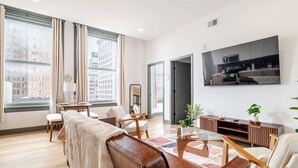 Un salon moderne comprenant un canapé, des fauteuils, une table basse, un téléviseur et une grande fenêtre donnant sur la ville.