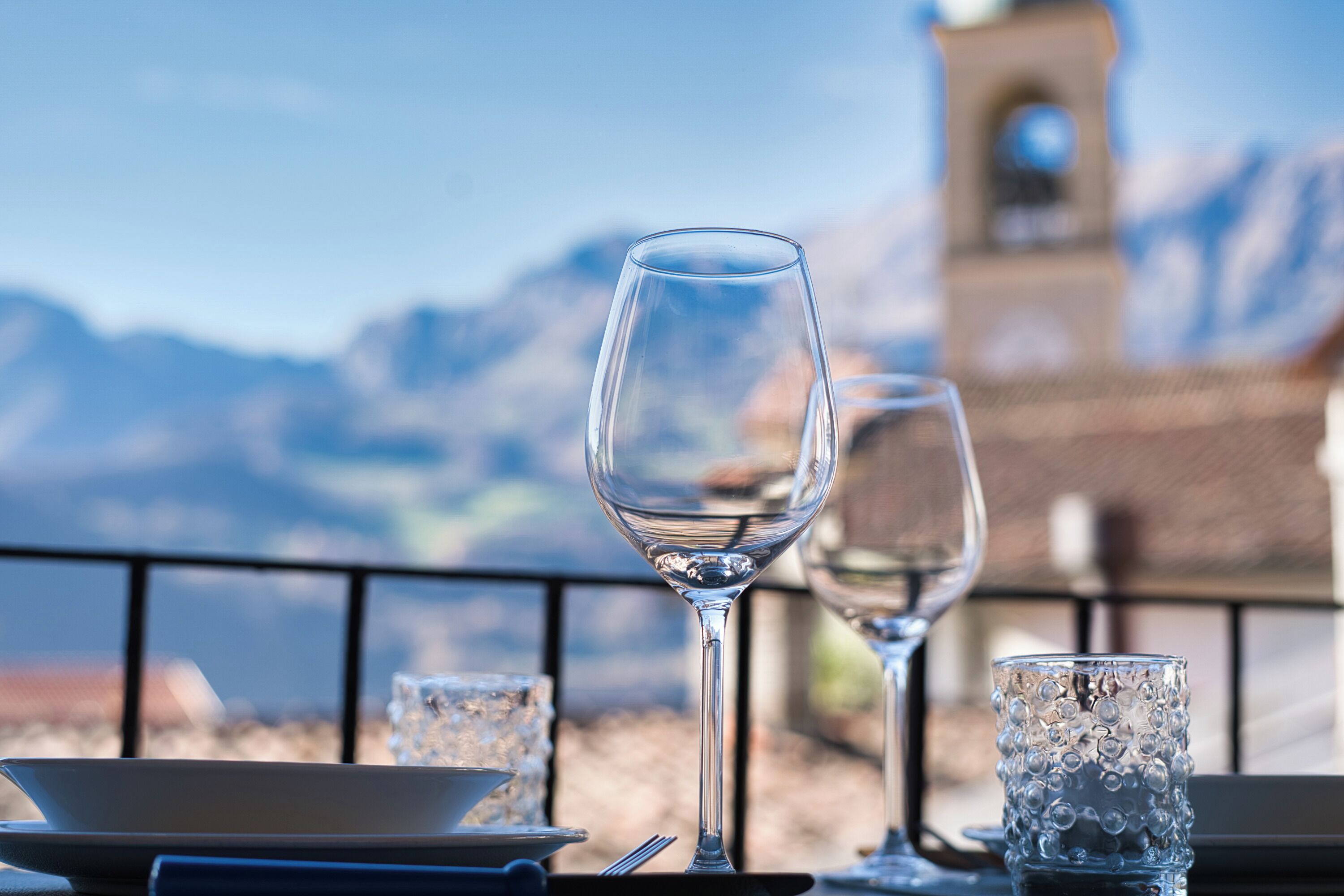 Ein Esstisch mit einem Weinglas und einem Glas Wasser, vor einer Bergkulisse mit einem Glockenturm.