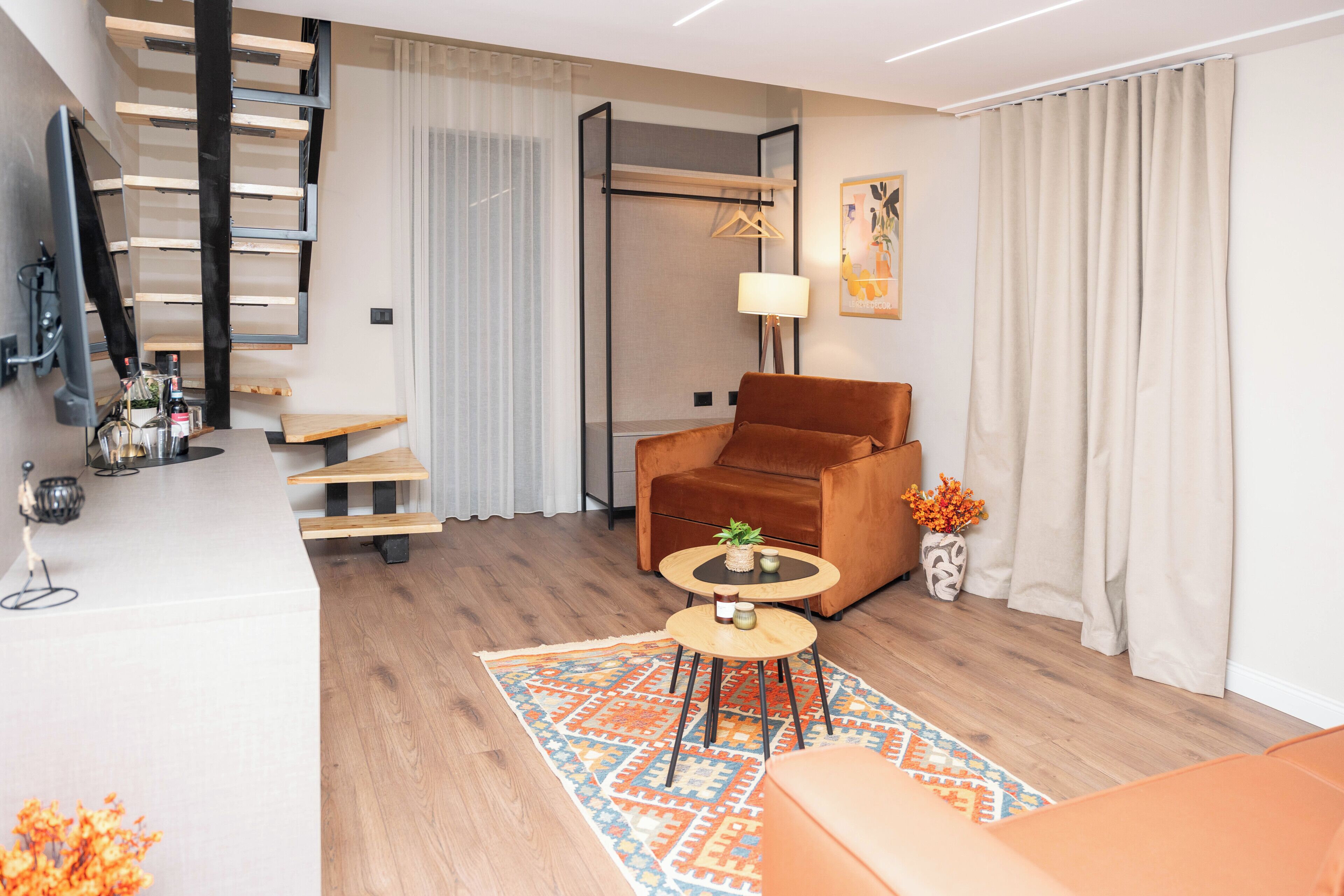 A modern living room with a brown leather sofa, a small round coffee table, and a patterned rug. A TV is mounted on the wall, and there is a staircase with wooden steps.