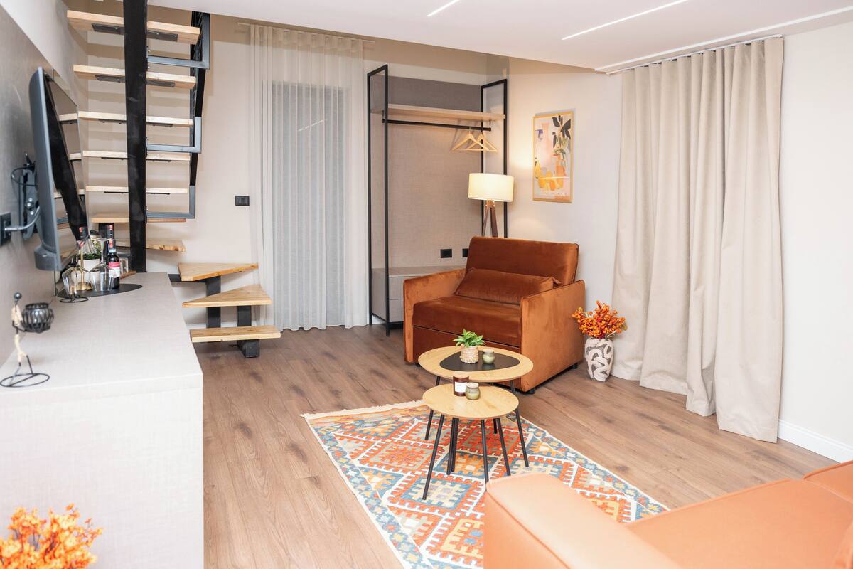 Una sala de estar moderna con un sofá de cuero marrón, una mesita de centro redonda y una alfombra estampada. Un televisor está montado en la pared, y hay una escalera con escalones de madera.