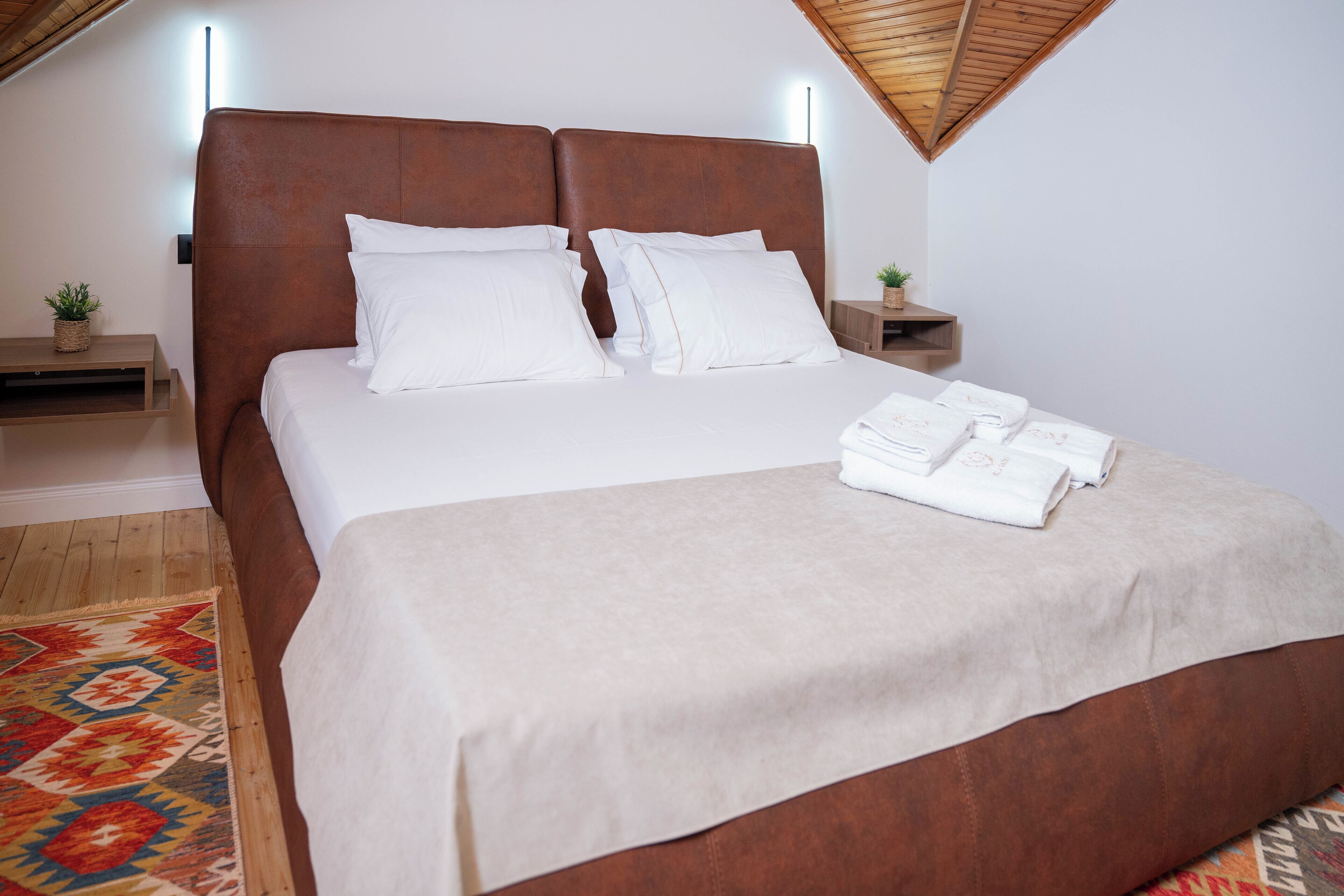 A neatly made bed with a brown headboard, white pillows, and a beige bedspread. There are two bedside tables with small plants and a wooden nightstand. A colorful rug is partially visible on the floor.
