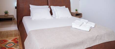 A neatly made bed with a brown headboard, white pillows, and a beige bedspread. There are two bedside tables with small plants and a wooden nightstand. A colorful rug is partially visible on the floor.
