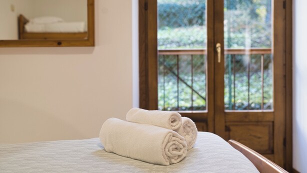 A neatly made bed with two rolled white towels, a wooden-framed window with a view of greenery, and a wooden door. - Lòbia in via Parrochiale - app. Pietra (P1) (Barzio)