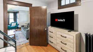 Ein Zimmer mit Holztür, einem an einer Kommode befestigten Fernseher, einer Treppe und Blick in einen weiteren Raum mit Sofa und Fenster.