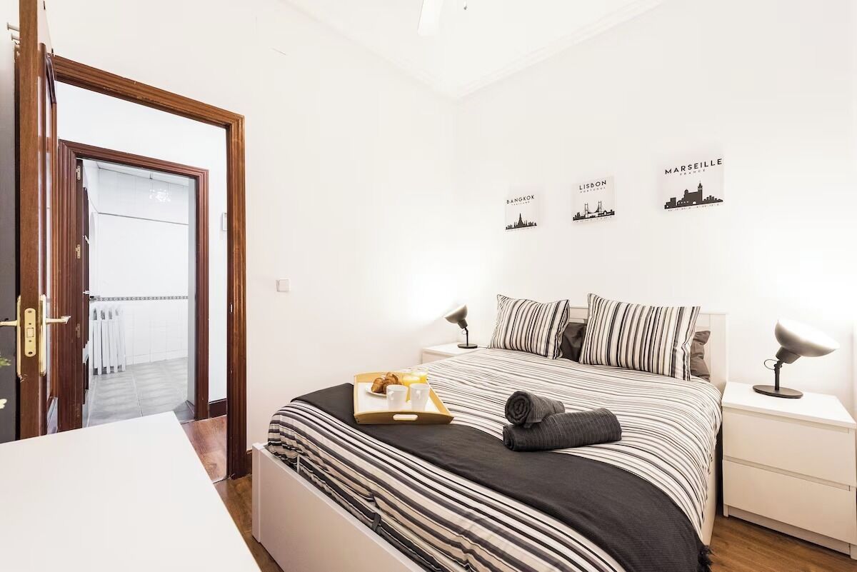A hotel room with a bed, a bedside table, a nightstand, a tray with breakfast items, and wall art depicting cityscapes.