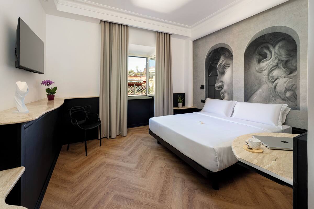 Una camera d'albergo con un letto, una scrivania, una sedia, una televisione e una finestra con tende.