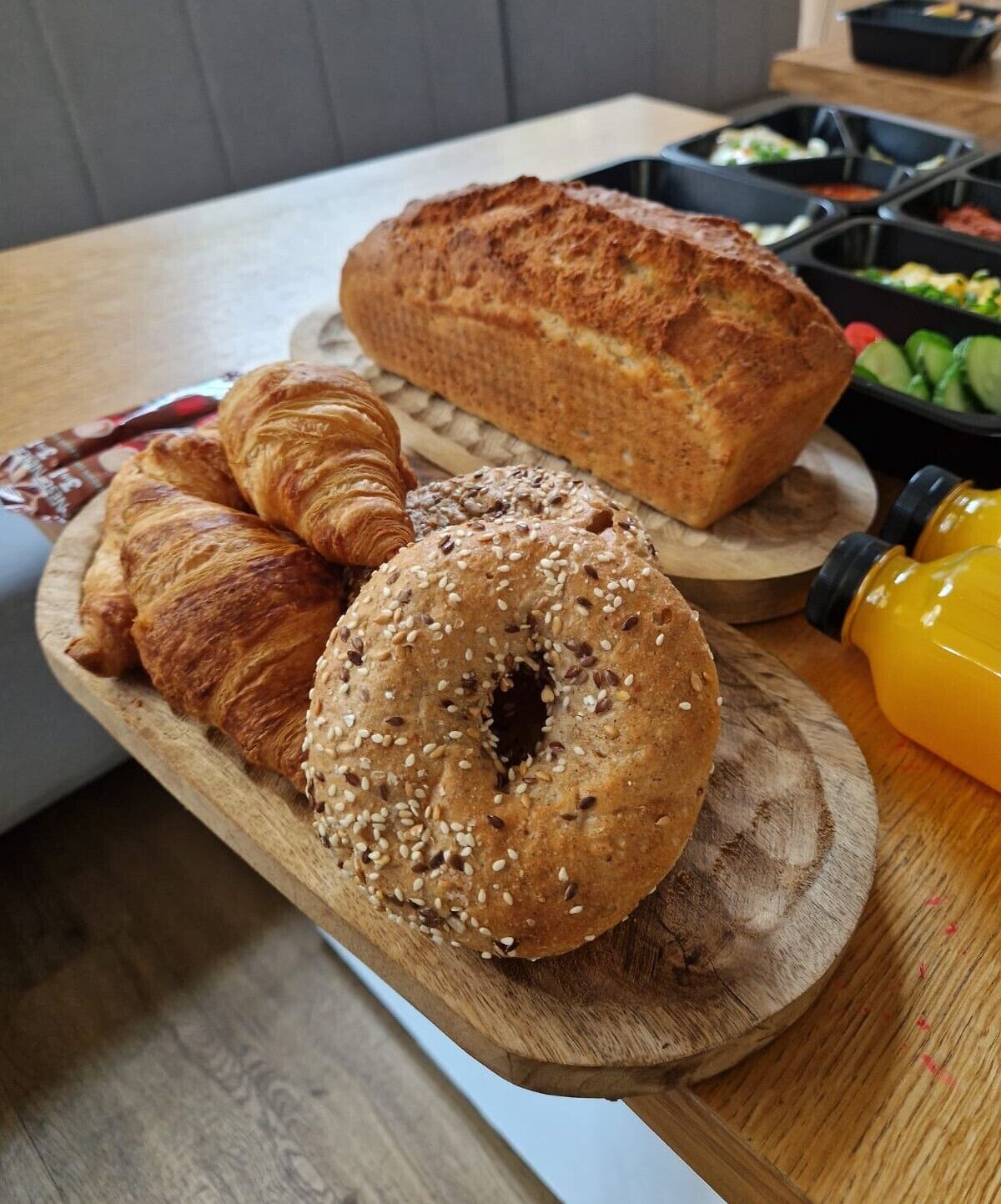 A wooden serving board with a croissant, a bagel, and a loaf of bread.