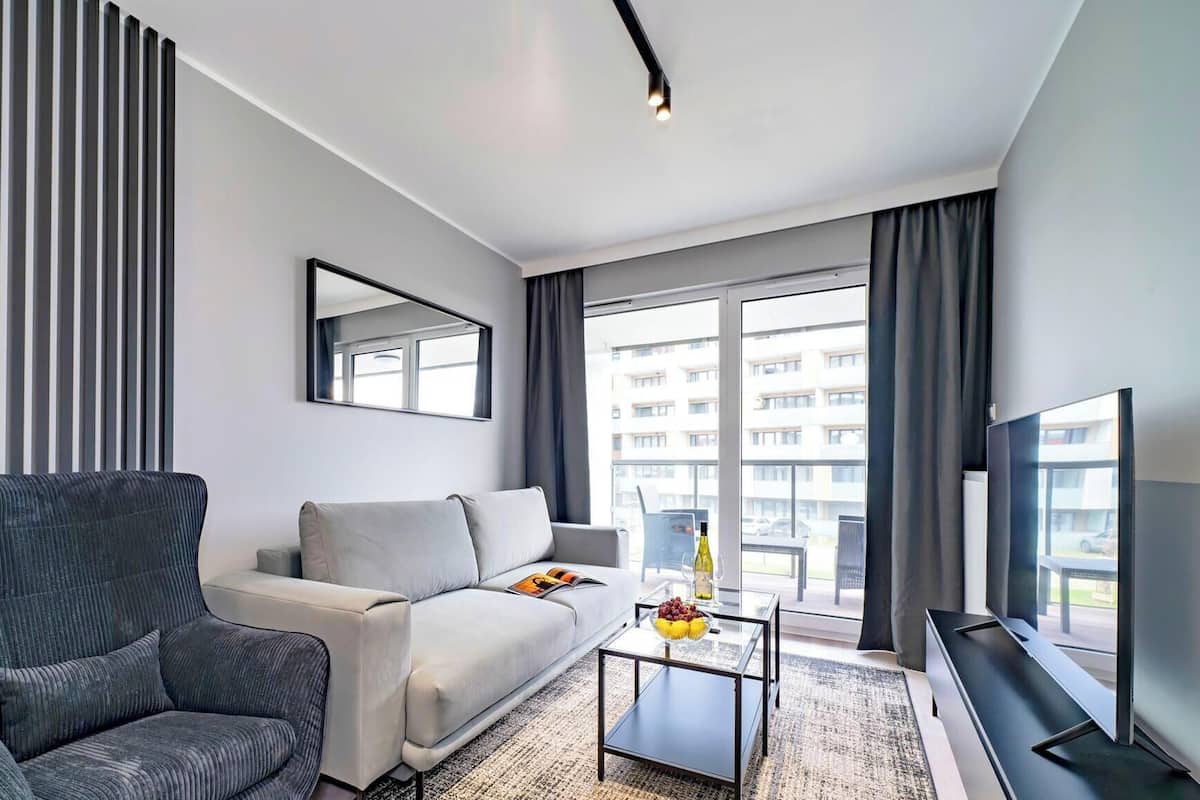 A modern living room with a grey sofa, a dark armchair, a coffee table with a magazine and a bottle, and a large window with a view of buildings.