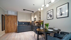 A modern kitchen with dark cabinets, a wooden door, and a dining area with a round table and chairs. - BalticON Apartments Kasprowicza (Kolobrzeg)