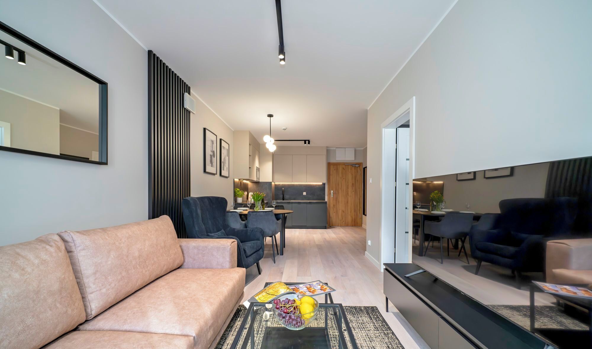 A modern living room with a sofa, chairs, and a coffee table with fruit. There is a kitchen in the background with a dining area.