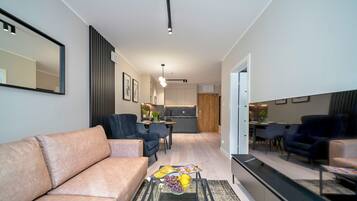 A modern living room with a sofa, chairs, and a coffee table with fruit. There is a kitchen in the background with a dining area.
