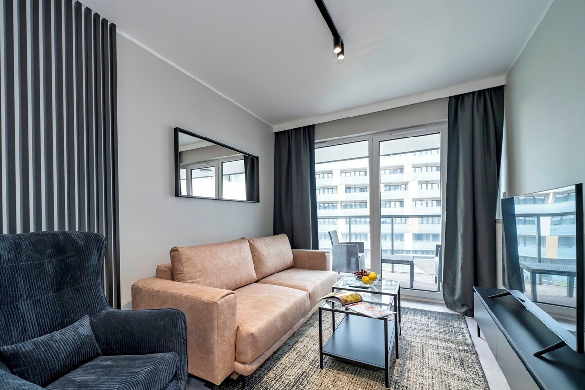 A modern living room with a sofa, a dark armchair, a coffee table, and a large window with a view of buildings.