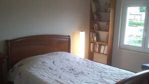 Une chambre avec un plafond en bois, un lit recouvert d’une couvre-lit à motifs, une table de chevet, une bibliothèque et une fenêtre donnant sur l’extérieur.