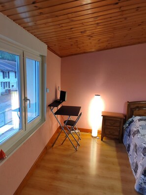 Een slaapkamer met een houten plafond, een bureau, een nachtkastje, een bed met een bloemenbedekking en een raam met uitzicht op gebouwen.