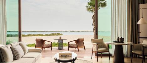 A room with a large window overlooking a beach, furnished with a sofa, chairs, and a table.