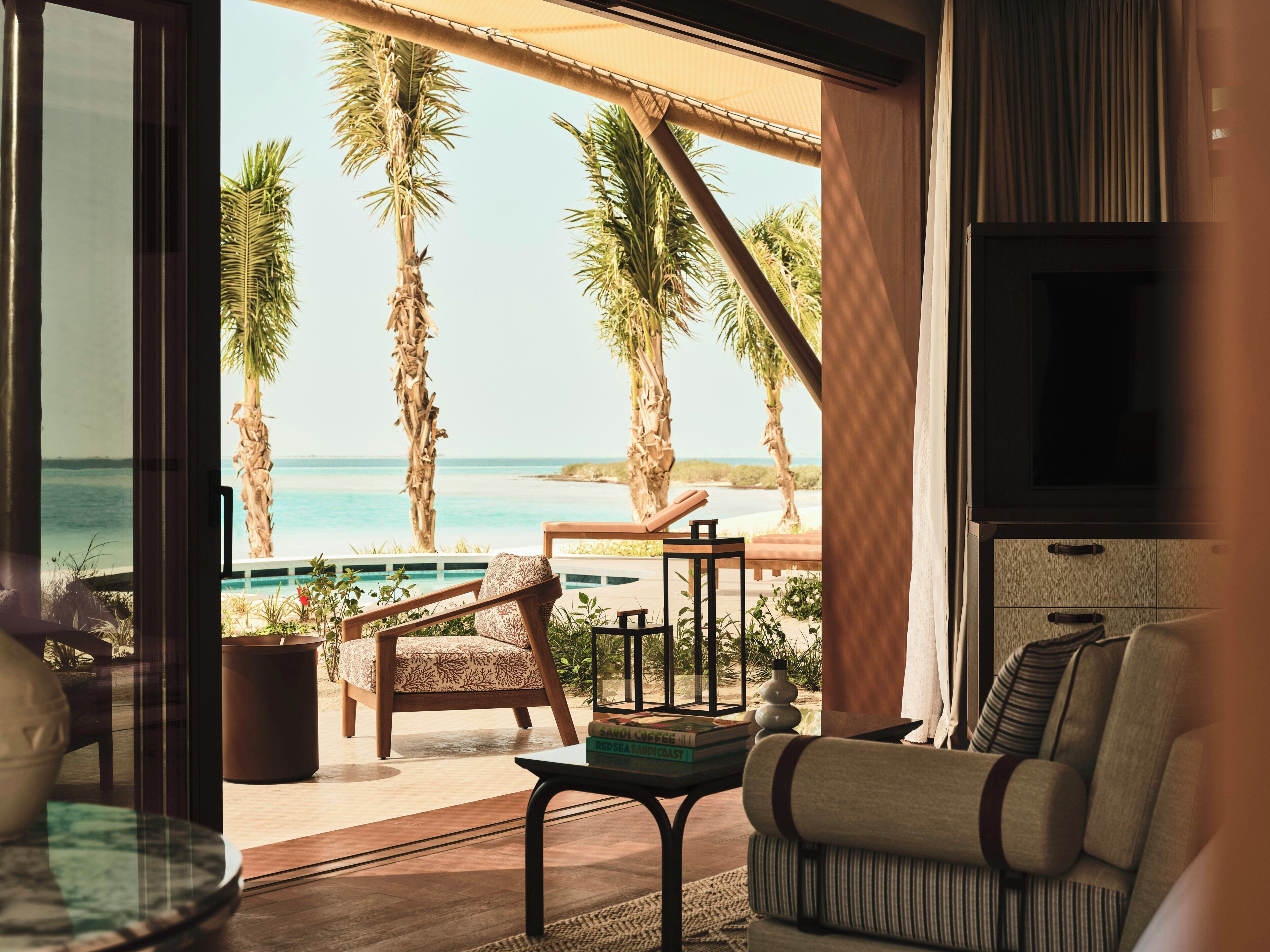 A room with a view of the beach, a pool, and palm trees. There is a sofa, a table, and a TV.