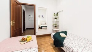 Quarto pequeno e arrumado com duas camas, uma porta de madeira, uma prateleira branca com garrafas e um quadro pendurado na parede.