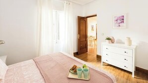 Quarto com cama, mesa de cabeceira com uma bandeja de croissants e uma xícara, cômoda branca e porta que dá para uma sala de jantar.
