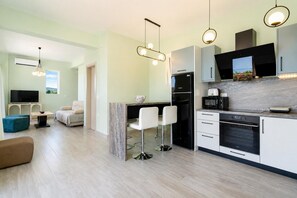 A modern kitchen with a built-in oven, a dining area with a bar stool, and a living room with a sofa. - Villa Aglaia - Two Bedroom Villa, Sleeps 4 (Mparmpati)