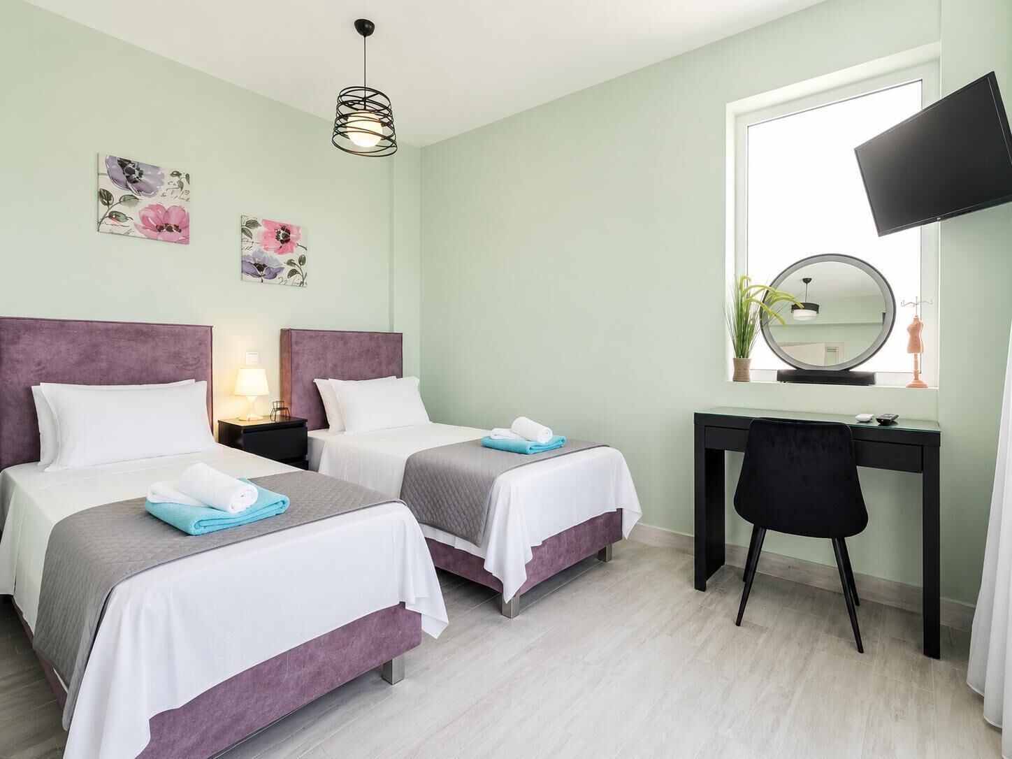 Quarto de hotel com duas camas, uma escrivaninha, uma televisão e uma janela com uma planta.