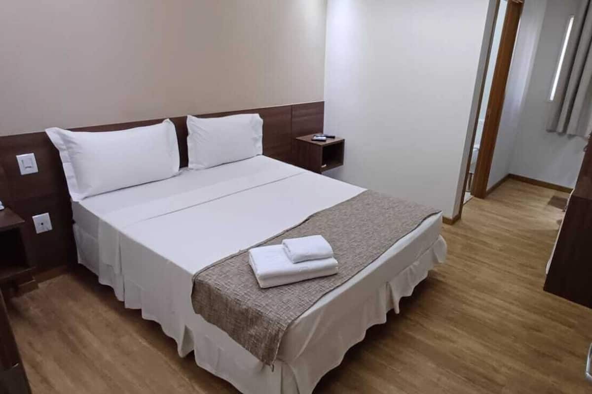 Una cama bien hecha con sábanas blancas y una toalla doblada, una mesita de noche con una lámpara y un suelo de madera.