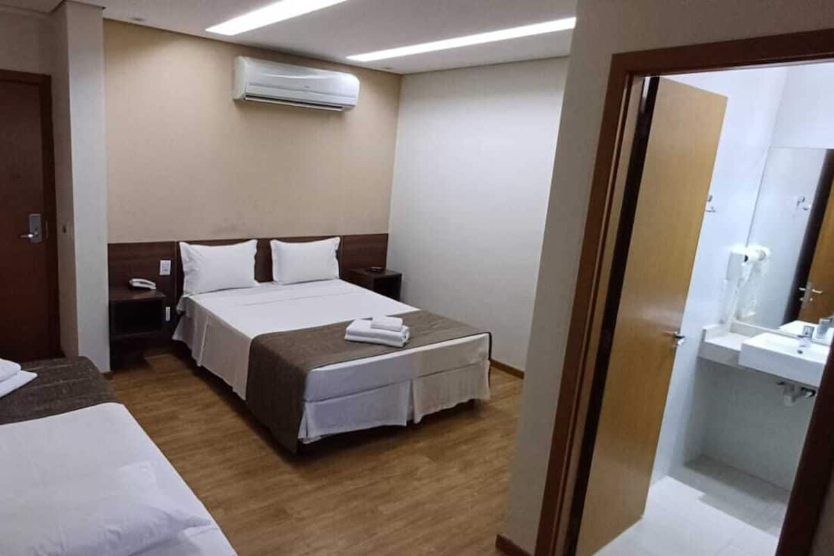 Habitación de hotel con dos camas, una mesita de noche con lámpara, aire acondicionado y baño con lavamanos y espejo.