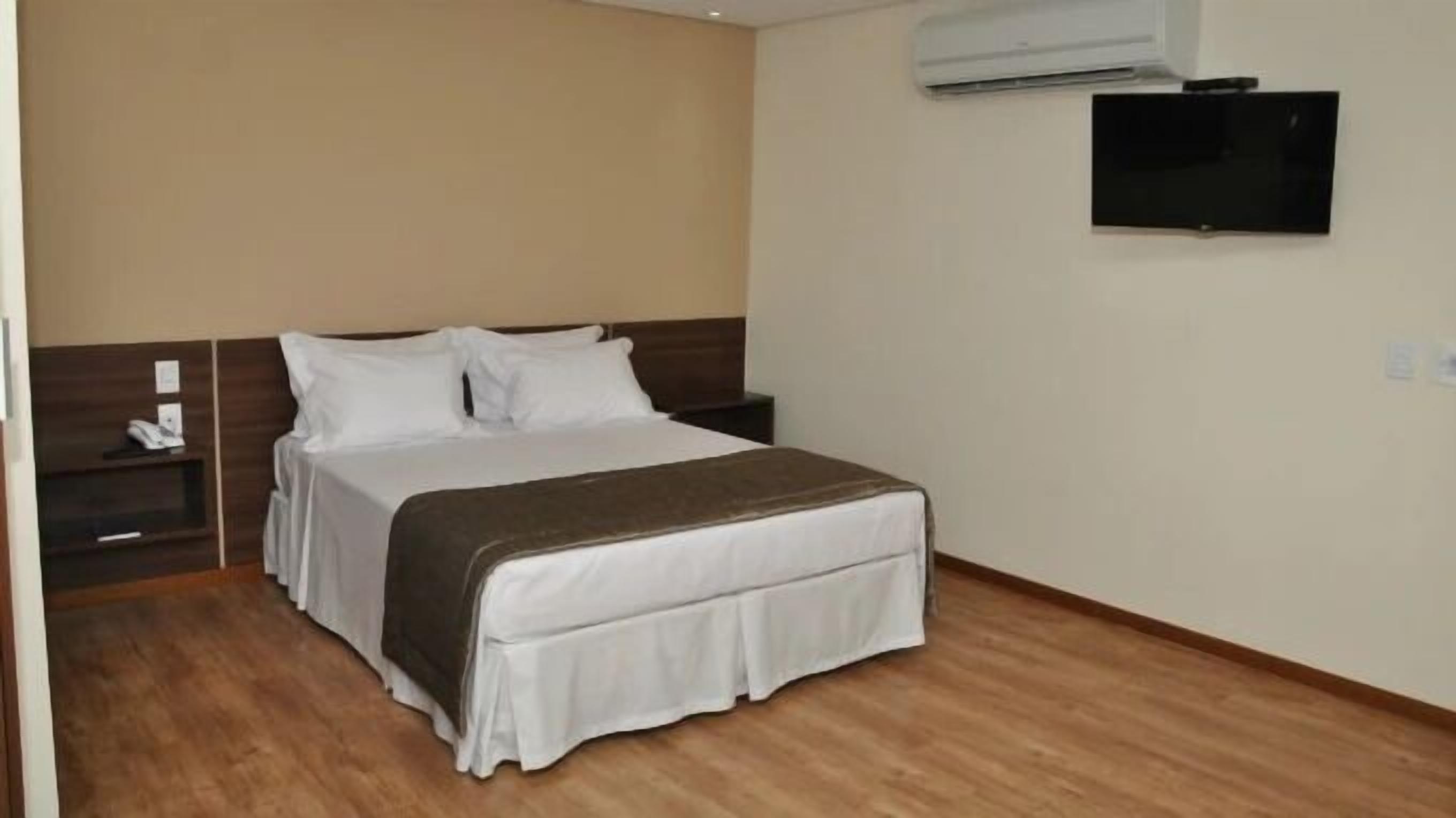 A hotel room with a bed, a nightstand, a TV, and an air conditioning unit.