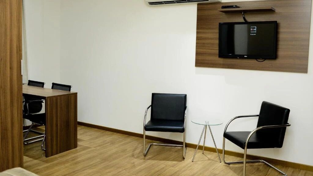 A conference room with a wooden desk, two black chairs, a small glass table, and a mounted flat-screen TV.