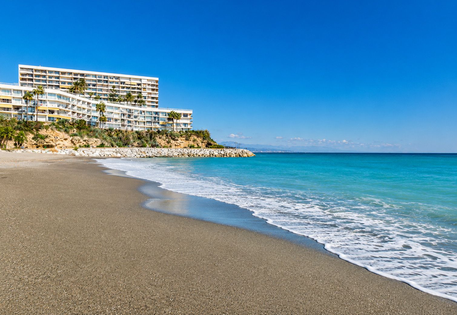 Un hôtel situé en bord de mer, doté de nombreux balcons, d’une plage de sable et offrant une vue sur un ciel bleu azur.