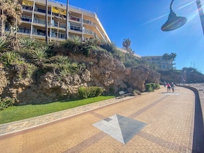 A coastal promenade with a paved walkway, a rocky cliff with vegetation, and a multi-story building with balconies. - Balcony overlooking the sea - Sea view (Torremolinos)