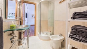 Un baño con cabina de ducha de vidrio, un lavamanos con repisa de vidrio, un bidé y un perchero con toallas dobladas.