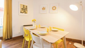 Un comedor con una mesa de madera preparada para cuatro personas, sillas amarillas y un jarrón con flores amarillas. Hay cuadros en la pared y una lámpara de pie.