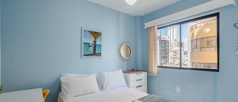 A bedroom with a bed, a desk, a chair, a window with a city view, and a ceiling fan.