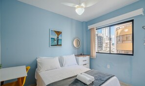 Un dormitorio con cama, escritorio, silla, ventana y vista a la ciudad, además de un ventilador de techo.