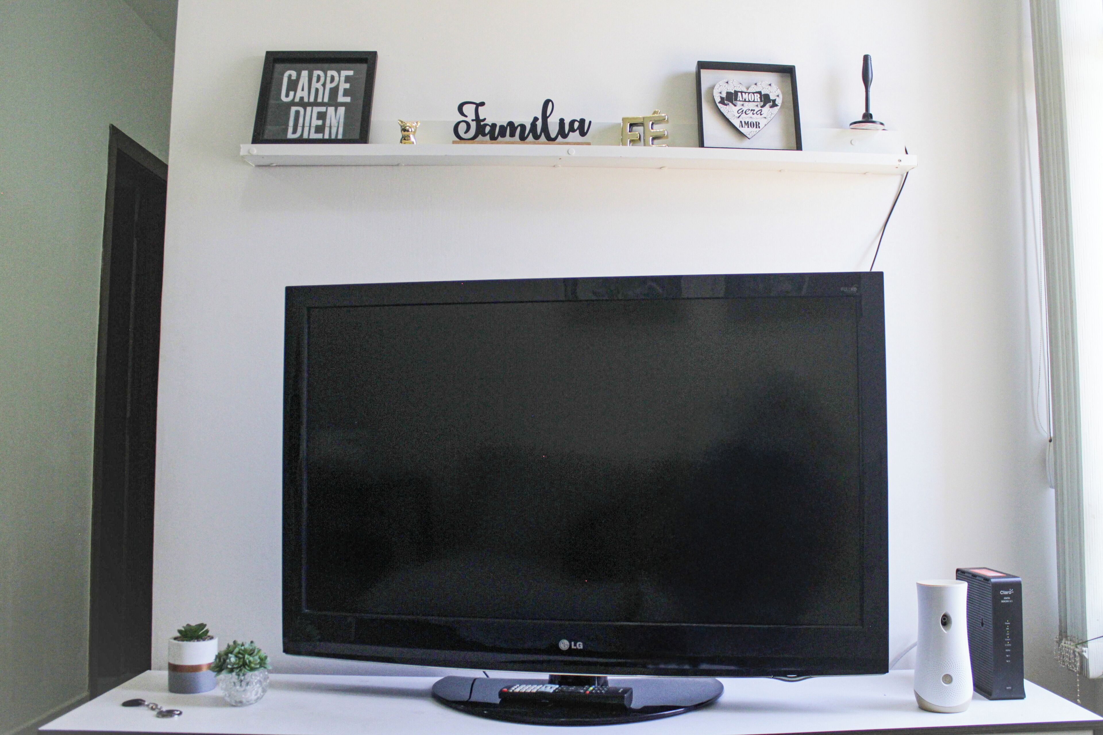 Una TV a schermo piatto su un supporto bianco, con un telecomando, una piccola pianta e un altoparlante poggiati su una scrivania bianca.