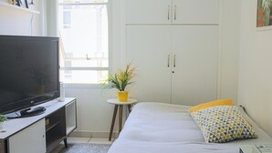 A bedroom with a bed, a TV on a stand, a small table with a plant, and a wardrobe. - Apartamento Toscana (Brasília)
