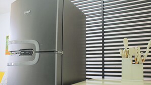 A modern kitchen with a stainless steel refrigerator, a white countertop, and a window with horizontal blinds. - Apartamento Toscana (Brasília)